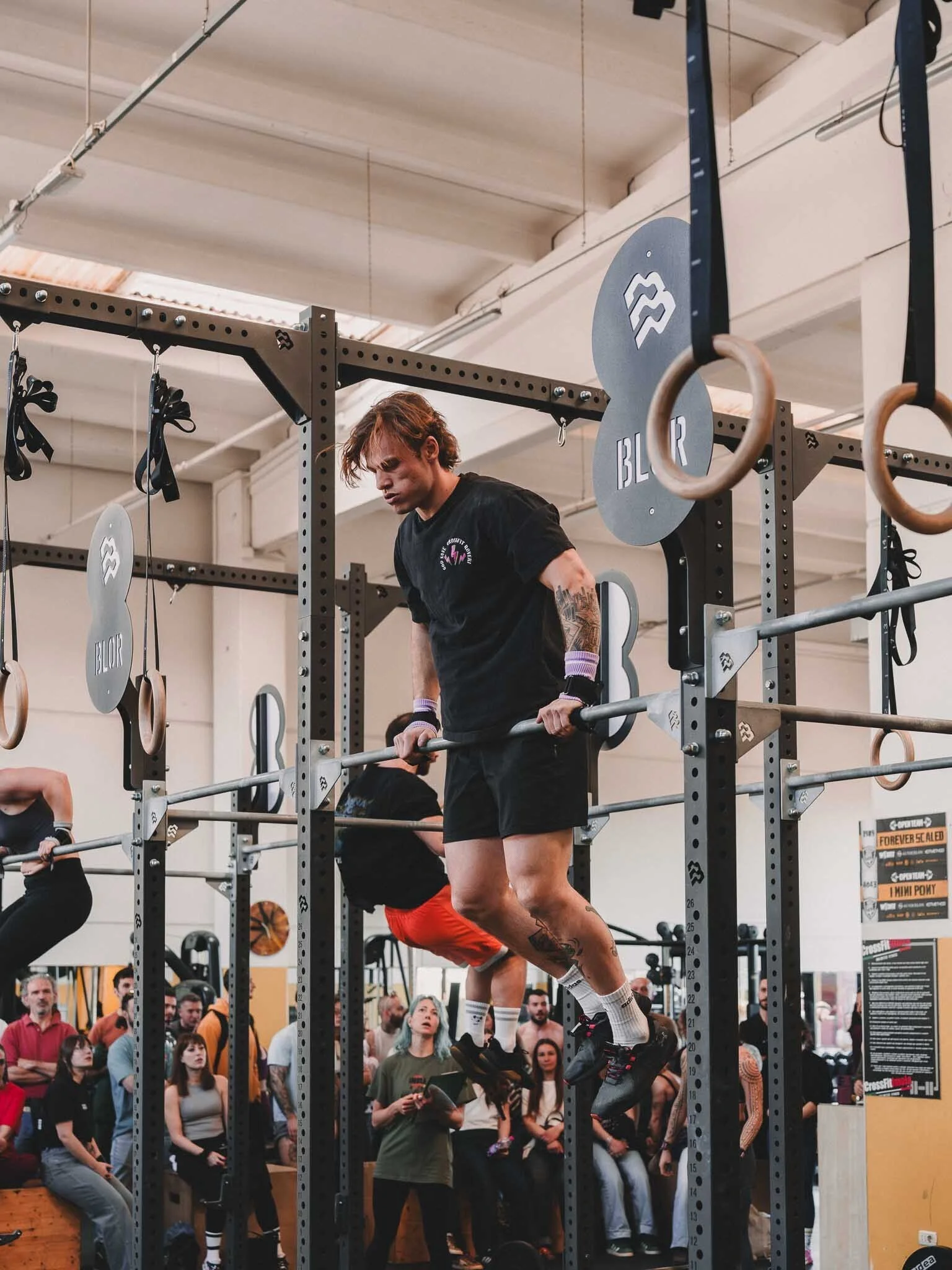 Uomo in allenamento di sollevamento pesi in palestra, circondato da persone che osservano, con attrezzature per il CrossFit visibili.