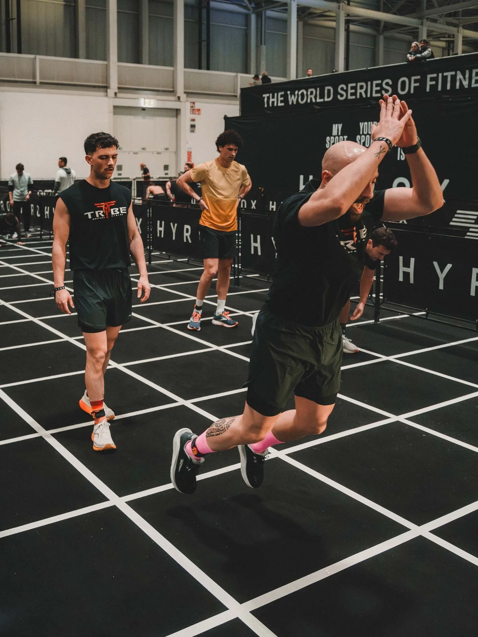 Un uomo che corre su una pista di atletica in un evento indoor, con altri atleti e persone sullo sfondo.