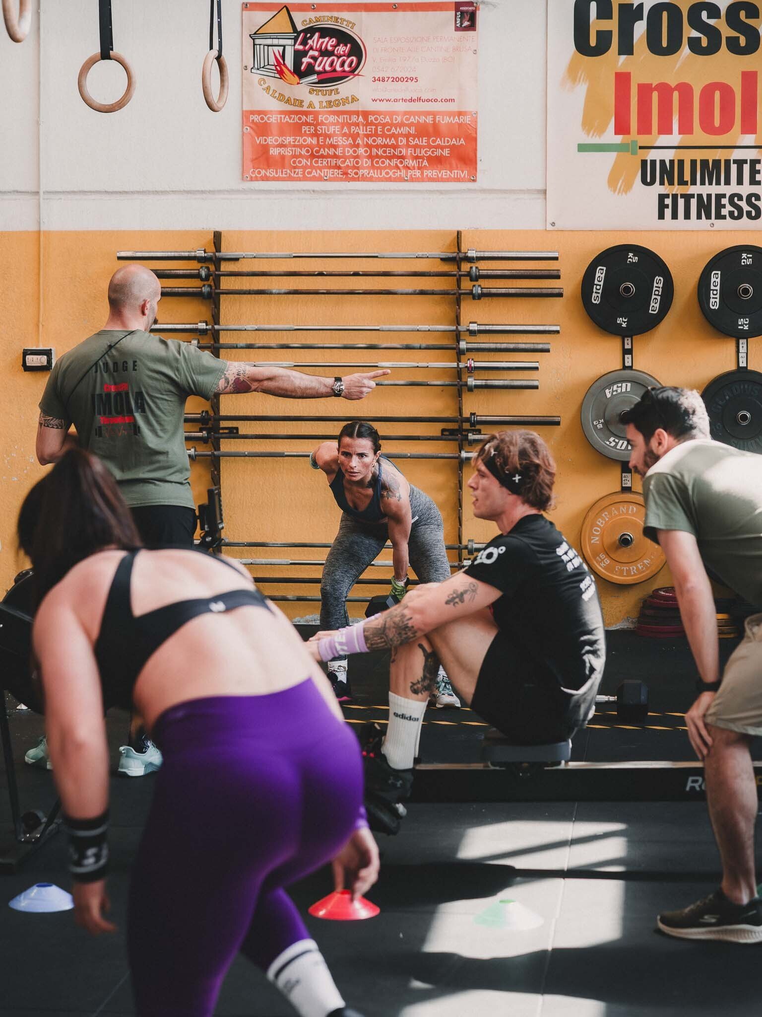 Un insegnante di fitness o personal trainer sta dando indicazioni a un gruppo di persone durante una sessione di allenamento in palestra. La scena si svolge all'interno di una sala con attrezzature da palestra, come bilancieri e pesi appesi al muro.