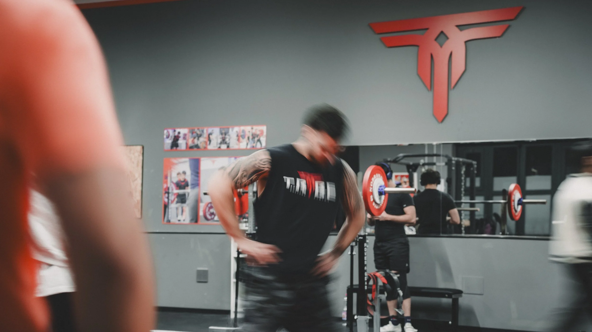 Persone che fanno esercizi in palestra, con attrezzature per il sollevamento pesi e uno sfondo con il logo di T-Fitness.