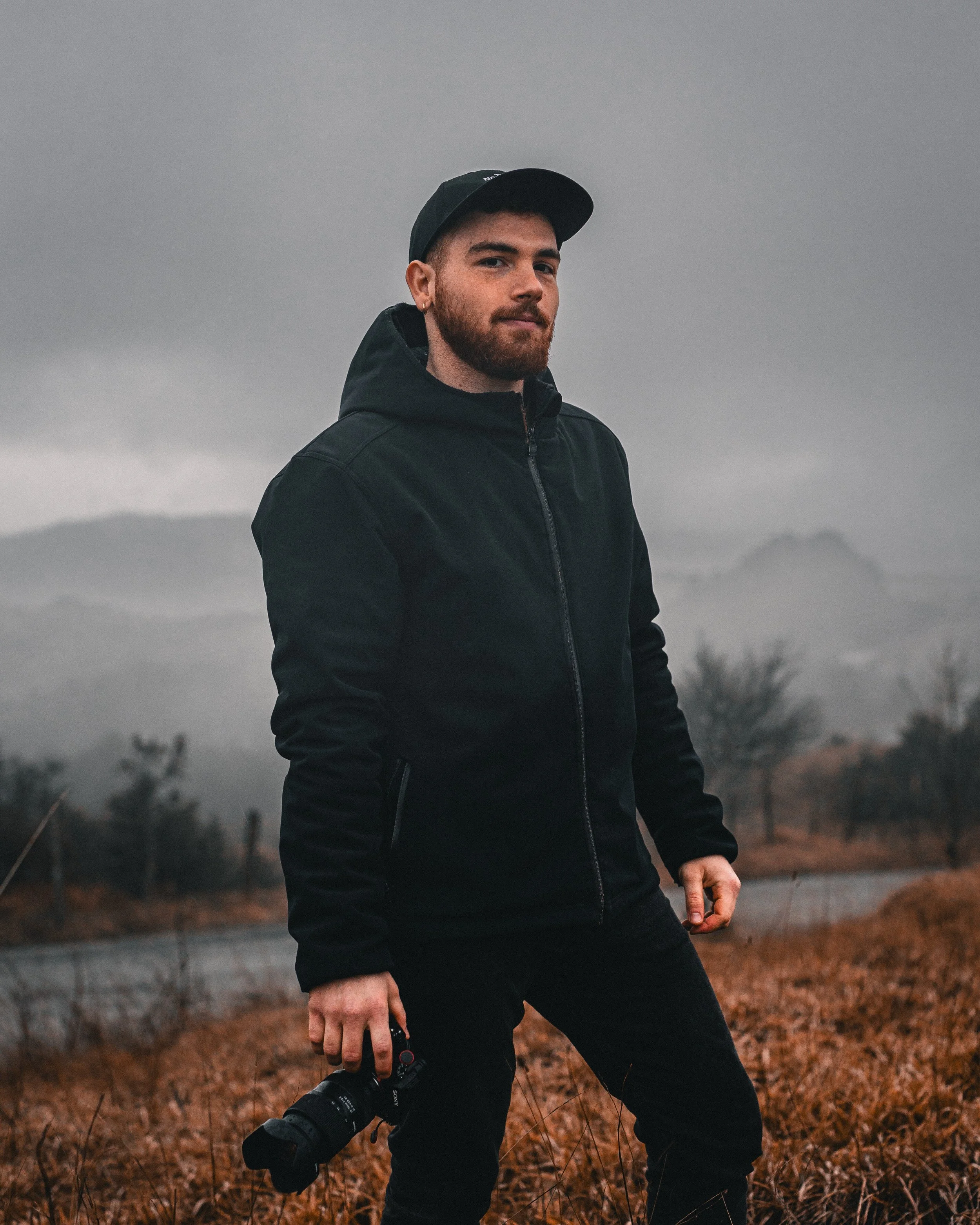 Uomo con cappello e giacca nera, tiene una fotocamera, sullo sfondo un paesaggio brumoso con montagne e alberi spogli.