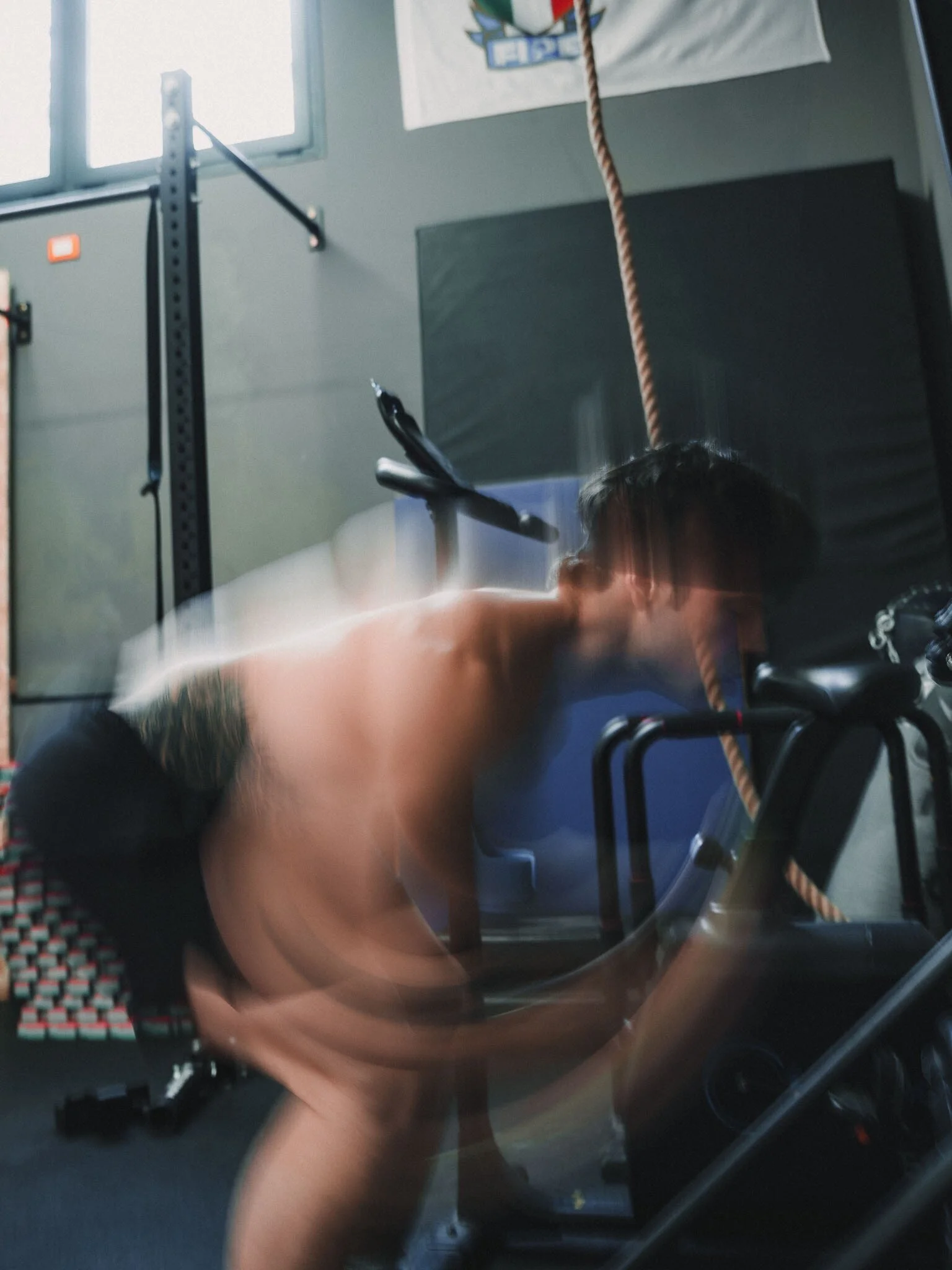 Persona nuda con capelli corti che si allena con una sbarra in palestra, di fronte a una parete con poster e finestre.