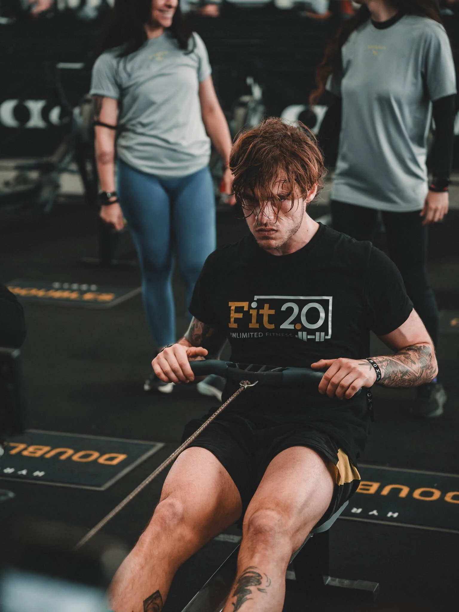 Un uomo con tatuaggi si sta allenando alla sbarra mentre due donne in tute sportive lo osservano in una palestra.