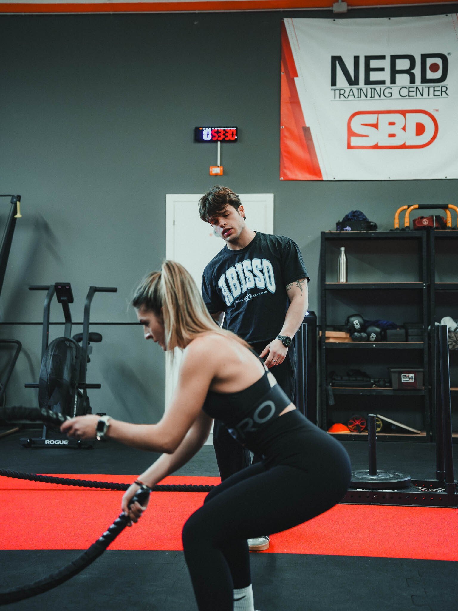 Una donna si allena con le kettlebell, mentre un uomo con maglietta nera e i pantaloni neri si guarda durante l'allenamento in palestra. Sul muro alle spalle c'è un orologio digitale e un banner con il logo "Nerd Training Center" e "SBD".