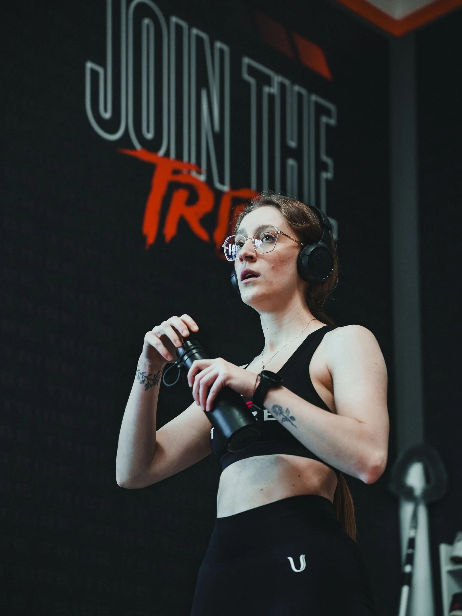 Donna con occhiali, capelli castani e tatuaggi, in palestra, tiene un kettlebell, sfondo con logo e attrezzi da palestra.