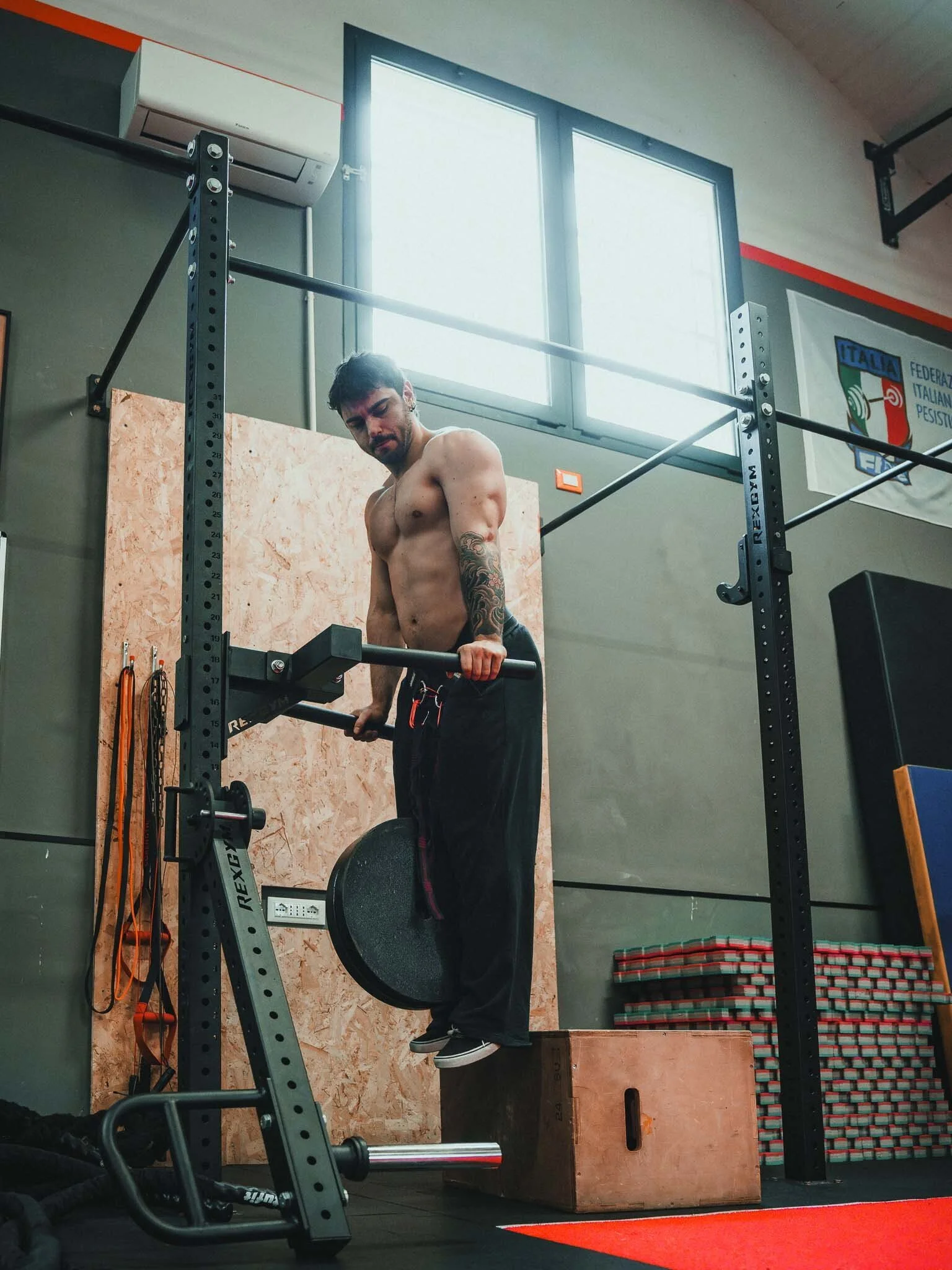 Uomo che si allena in palestra con una barra, senza maglietta, con una parete in legno dietro e una bandiera dell'Italia e della Federazione Italiana di Pesistica appese alla parete.