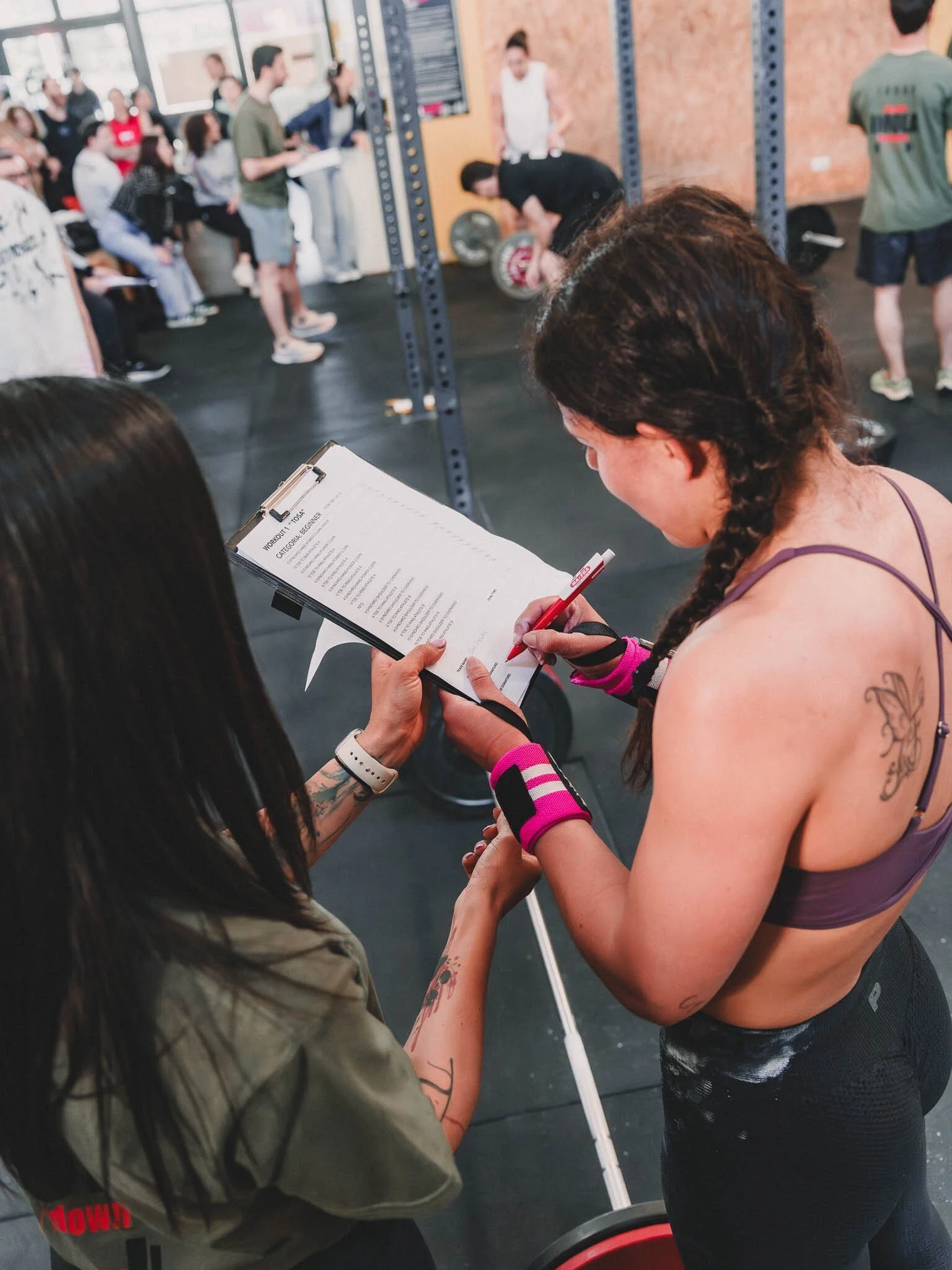 Una donna con tatuaggi stringe la mano di un'altra donna mentre tiene un blocco notes con un foglio, in un centro fitness. Sullo sfondo ci sono persone che eseguono esercizi e altre che aspettano, in un ambiente industriale con scaffalature e attrezz