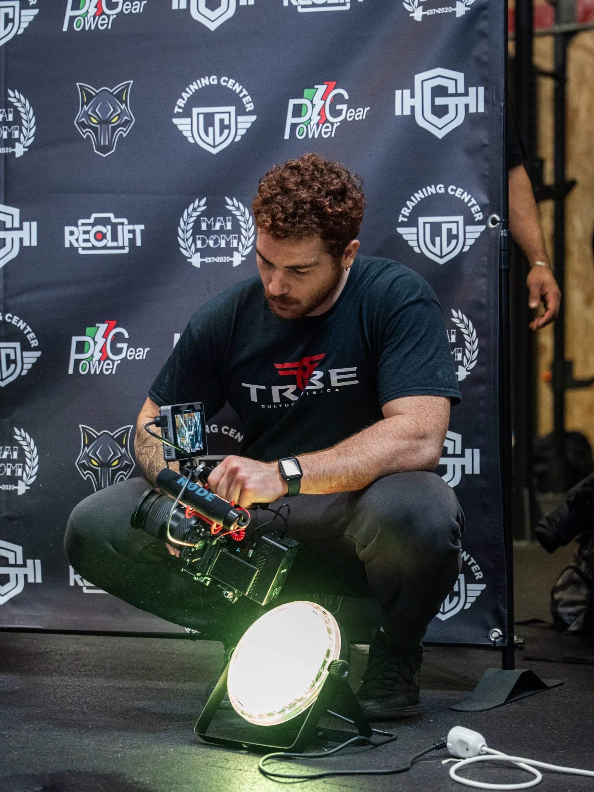 Un uomo con capelli ricci e barba, seduto su un pavimento nero, usa una videocamera con uno schermo esterno, illuminato da una luce circolare verde, contro uno sfondo con vari loghi e scritte di centri di allenamento.