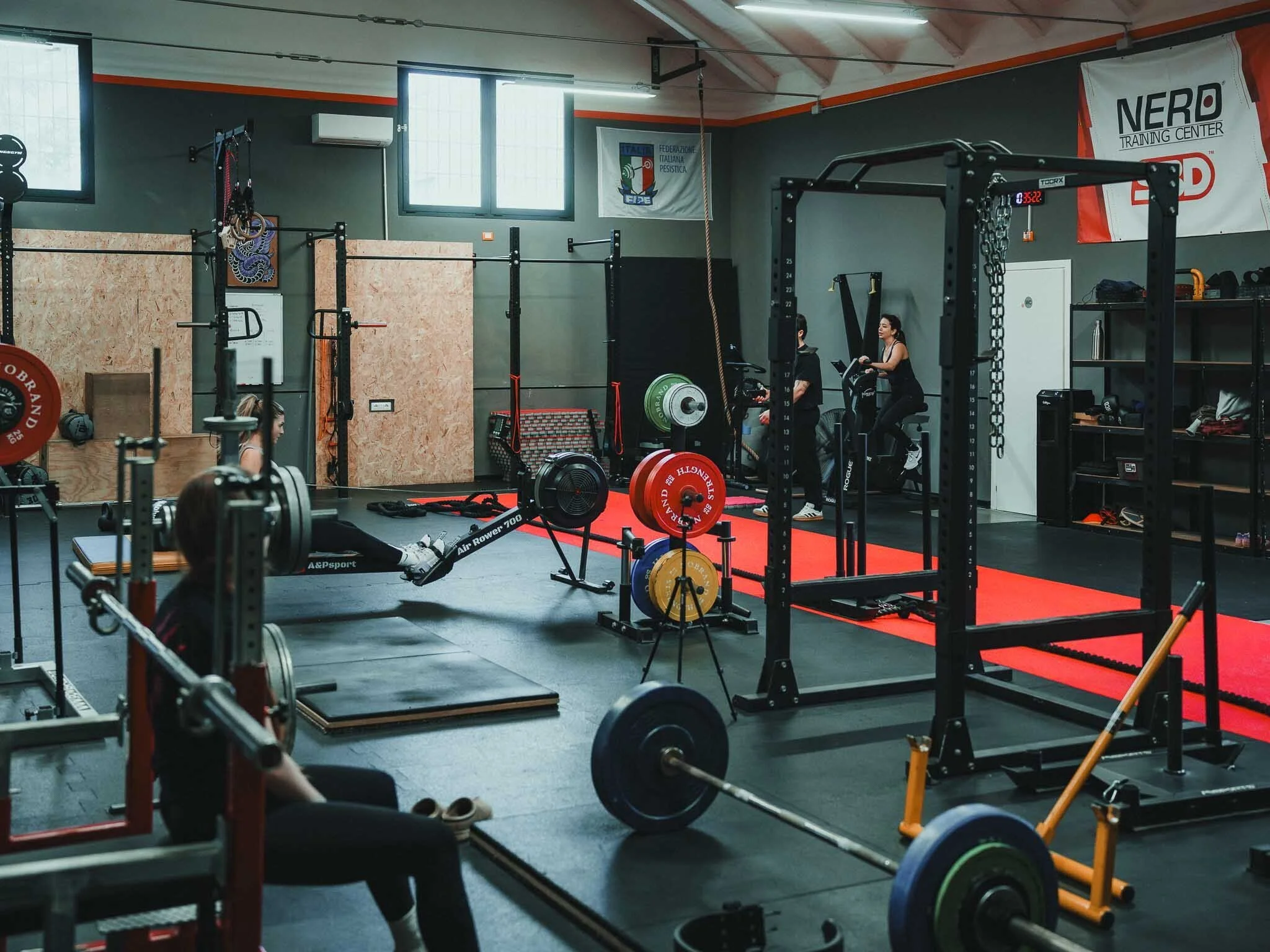 Interno di una palestra con varie attrezzature di allenamento, tra cui bilancieri, dischi pesi e macchinari, con alcune persone che si allenano.