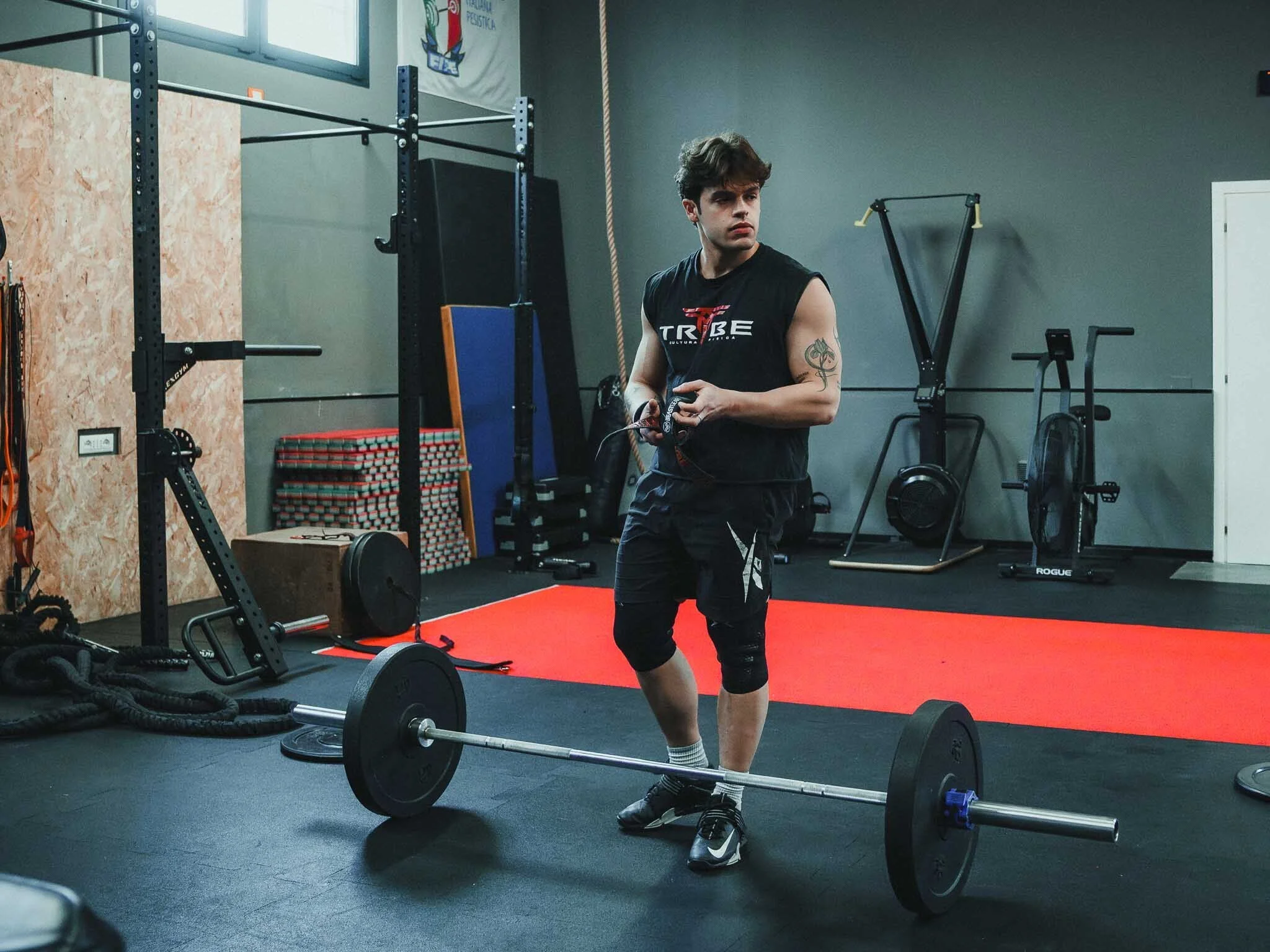 Un giovane uomo si allena in palestra, indossa una maglietta senza maniche con il logo TRIBE, e si sta preparando a fare sollevamento pesi con un bilanciere sul pavimento. La palestra ha attrezzature per il fitness, una parete con una bandiera e pare