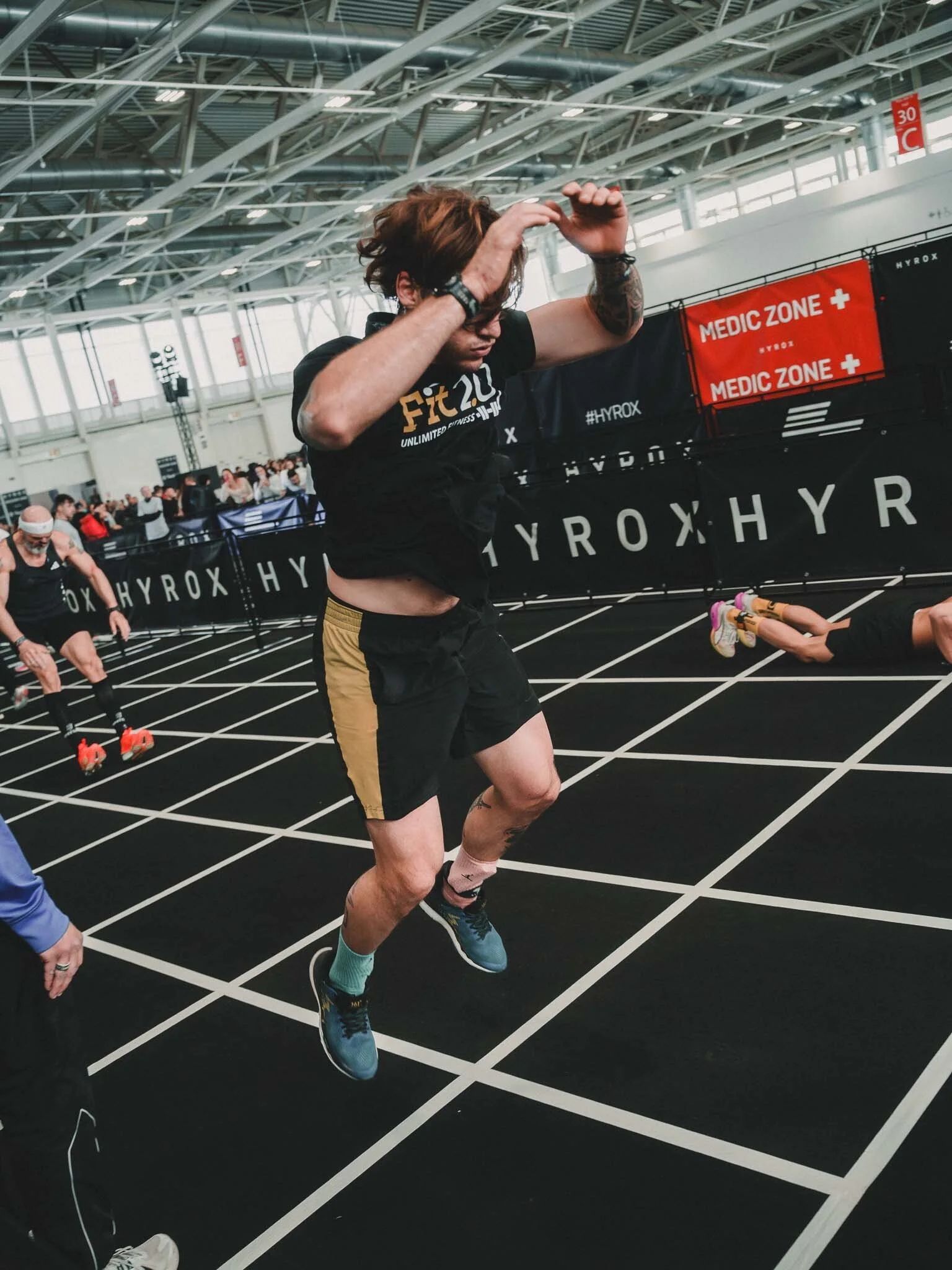 Uomo che corre su una pista indoor, partendo da una posizione di corsa, con persone intorno e segnali di emergenza e aree mediche sullo sfondo.