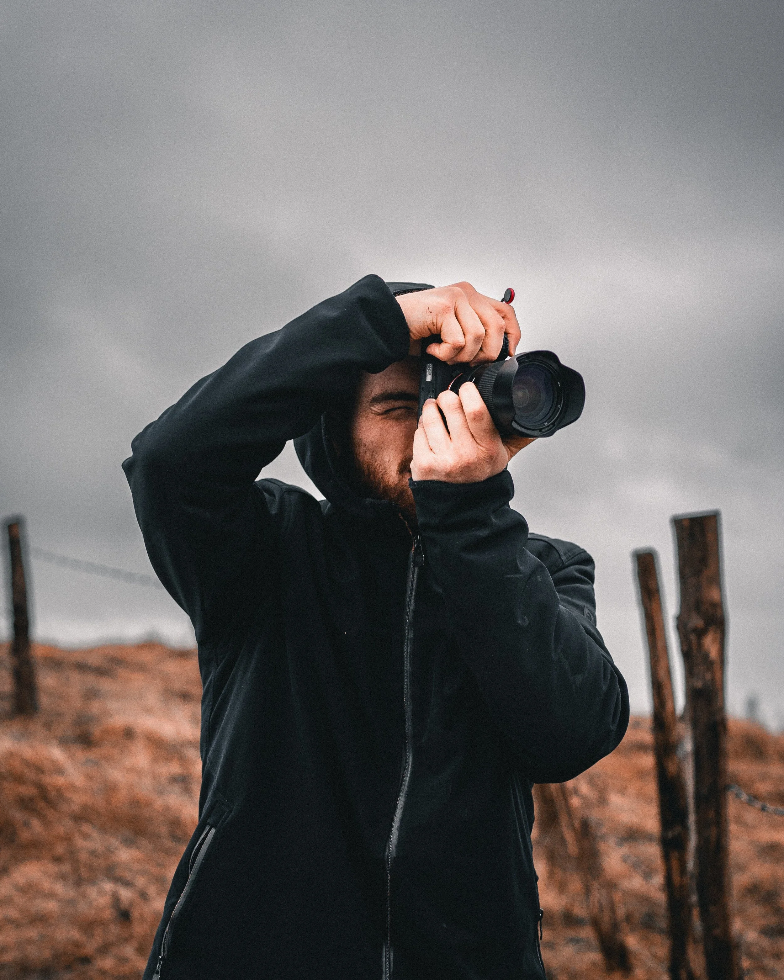 Un uomo indossa una giacca nera e fotografa con una fotocamera reflex in un ambiente all'aperto con cielo nuvoloso e recinzioni di legno sullo sfondo.