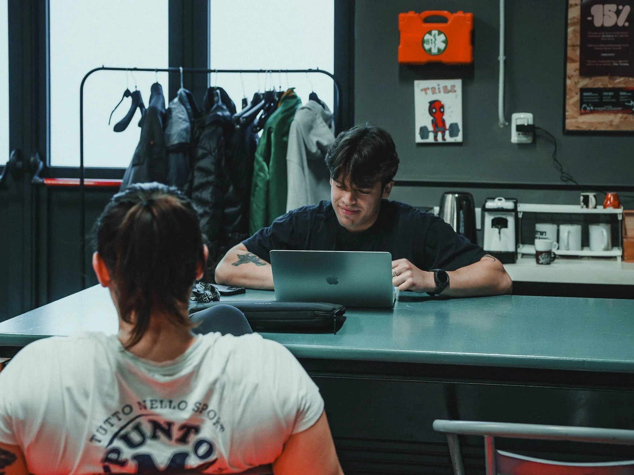 Due persone sedute a un tavolo di ufficio o caffetteria, una con cappello bianco e stampe colorate, l'altra con capelli corti e maglietta nera, una MacBook aperto davanti a lei. Sullo sfondo si vedono ganci con abbigliamento e una parete con poster e