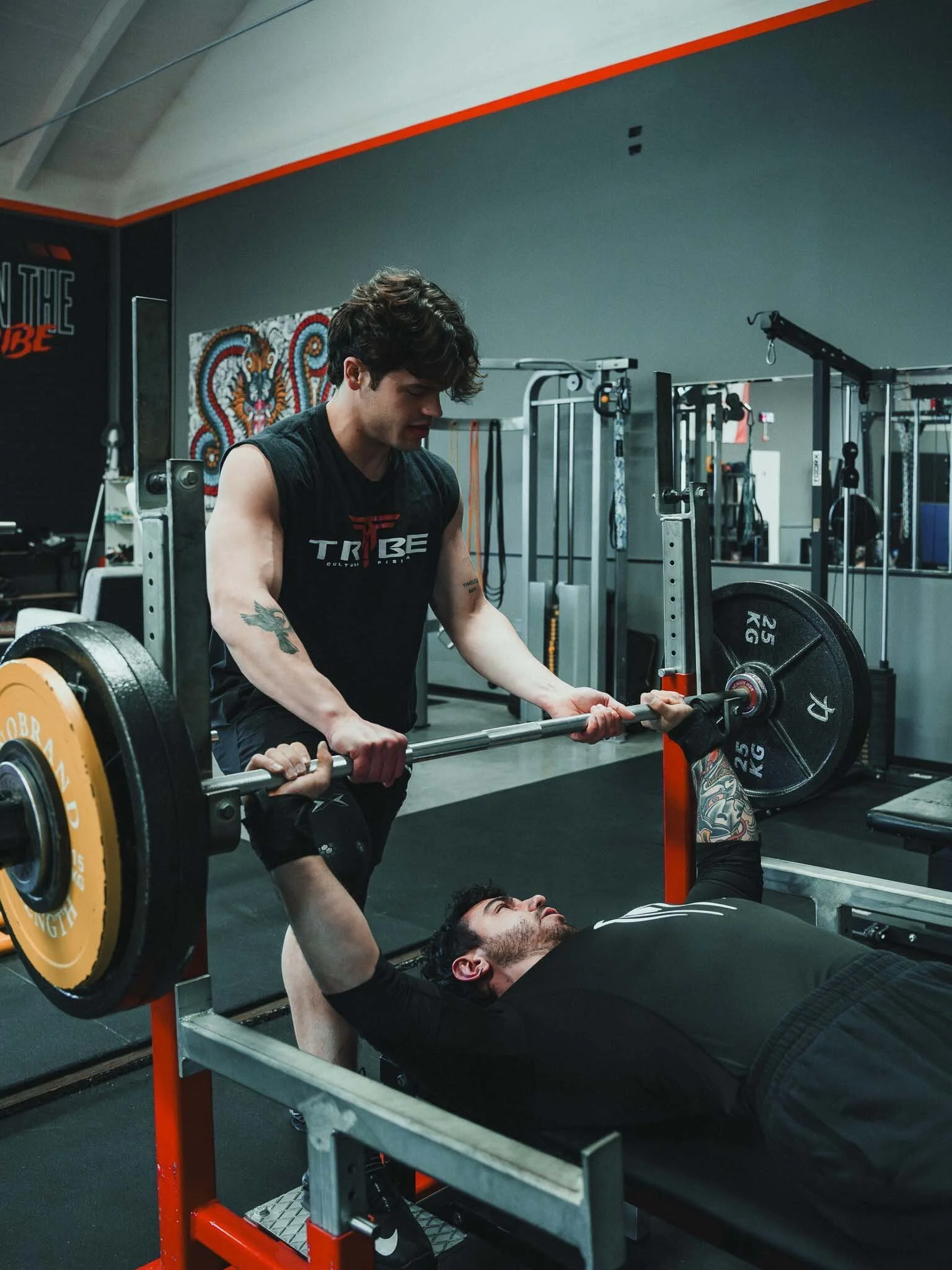 Un uomo sta sollevando un bilanciere durante l'allenamento, con un partner che lo assiste mentre è sdraiato su una panca nel palestra.