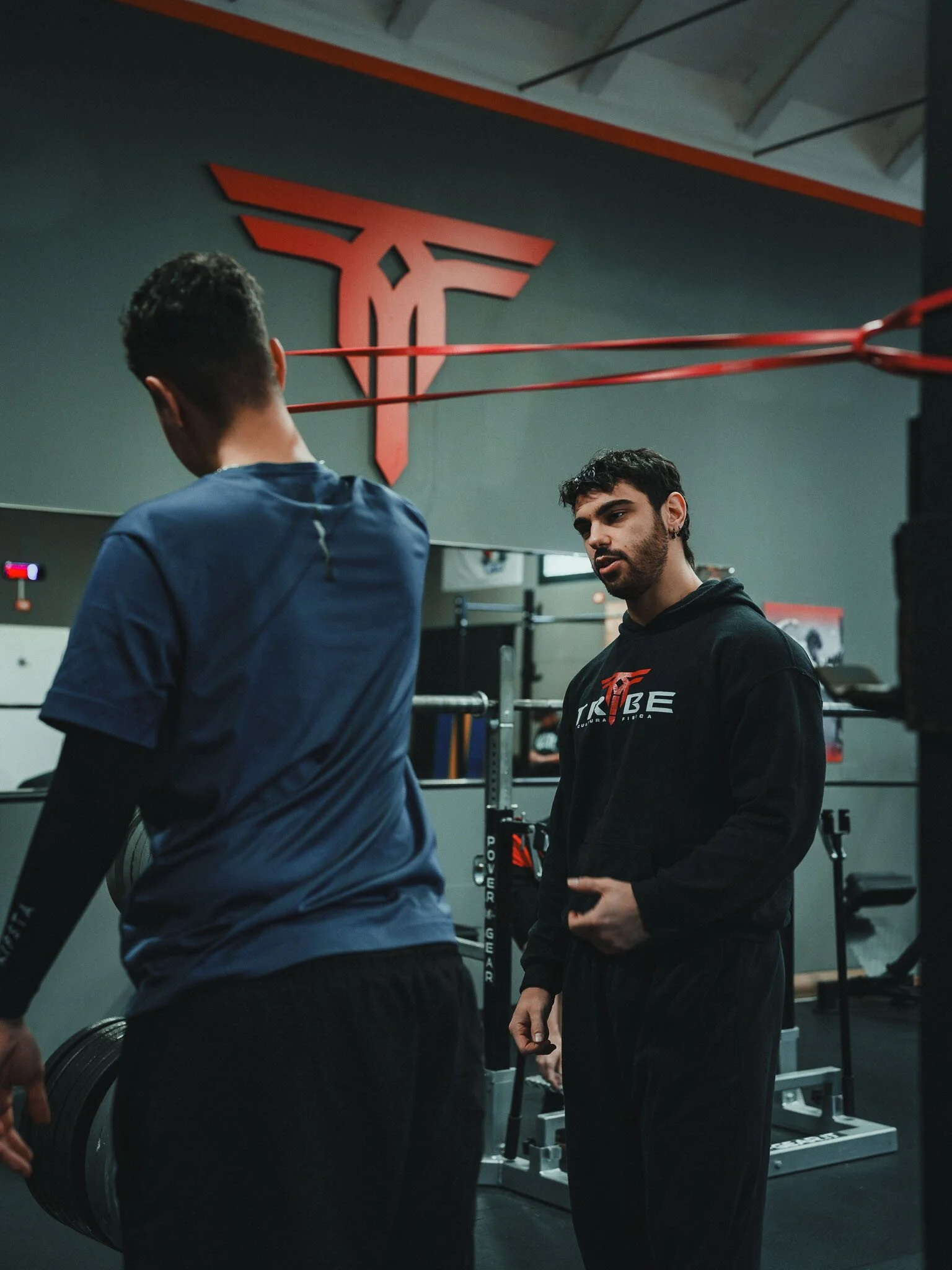 Due uomini in palestra, uno con una maglietta blu e l'altro con felpa nera con logo, che si allenano con manubri e pesi, in un ambiente con logo rosso di un'ala e un teschio sulla parete.