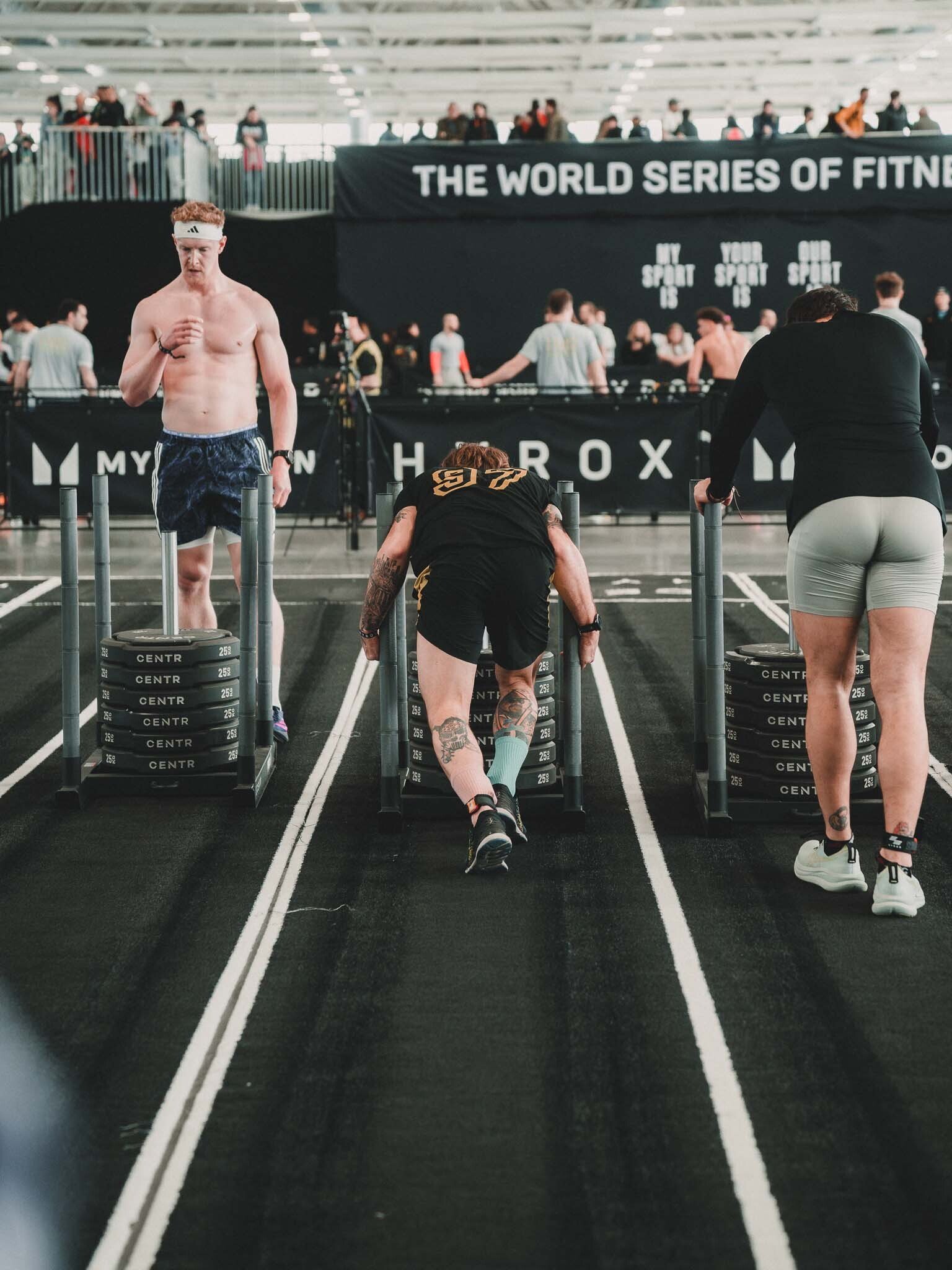 Atleti che si preparano per la competizione di CrossFit in un'area dedicata all'interno di una struttura sportiva, con uno sfondo che indica un evento di fitness internazionale.