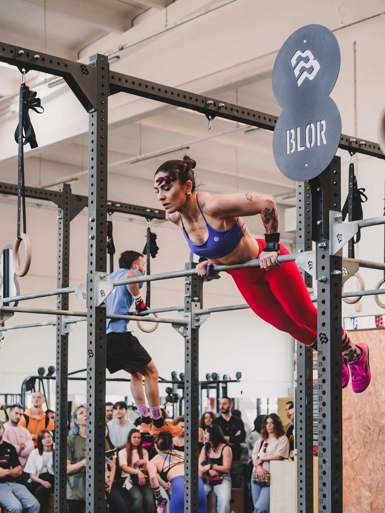 Una donna atletica si allena in una palestra di crossfit, facendo esercizi con una barra mentre un pubblico osserva, con vari attrezzi e una folla di spettatori sullo sfondo.