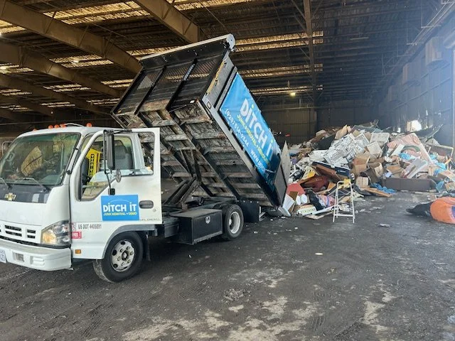 A small white trash removal truck with a blue Ditch It logo on the side is dumping a large pile of trash, including cardboard boxes, furniture, and debris, inside a warehouse or industrial space.