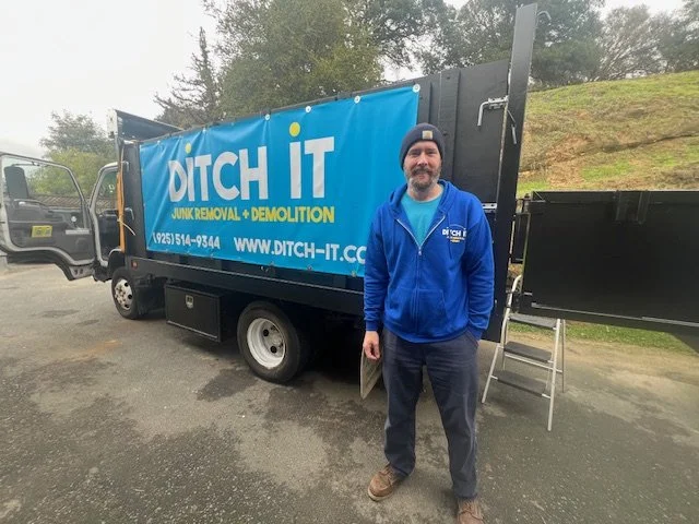 Man standing in front of a DITCH IT junk removal truck wearing a blue hoodie and a black beanie.