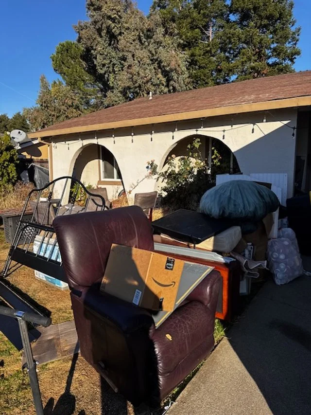 Outdoor scene with household items piled in front of a house, including a brown leather armchair, bedframe parts, various boxes, and other miscellaneous belongings. The house has arched windows and a sloped roof, surrounded by trees.