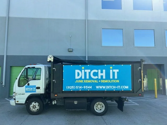 A small truck with a blue sign on the side that says "DITCH IT" with phone number and website, parked outside a building with gray walls and windows.