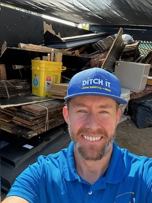 A man with a beard wearing a blue cap that says "Ditch It Junk Removal & Demo" and a blue polo shirt, smiling in front of a pile of scrap materials including wood, metal, and a yellow bucket.