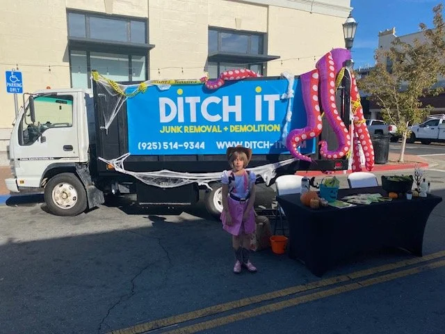 A decorated junk removal truck with a Halloween theme, featuring a large tentacle and cobwebs, parked on the street. A young girl in a costume stands in front of a table with pumpkins and promotional materials. A building with a handicap parking sign is in the background.