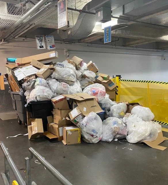 Overflowing trash bins filled with cardboard boxes and plastic bags in an underground parking garage.