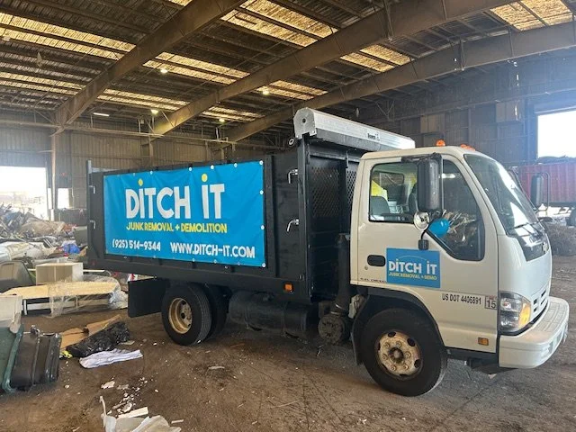A small white work truck with a blue advertisement for Ditch It, a junk removal and demolition company, parked inside a warehouse. The truck has a black storage container on the back and is surrounded by scattered debris and items on the ground.