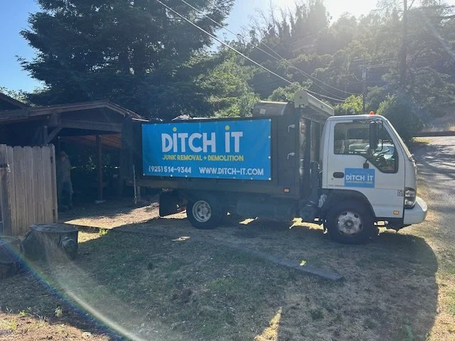 A small gray and blue truck with a large sign on its side reading 'DITCH IT Junk Removal & Demolition,' parked on a patch of grass near a wooden structure. The truck is on a residential street with trees and power lines in the background.
