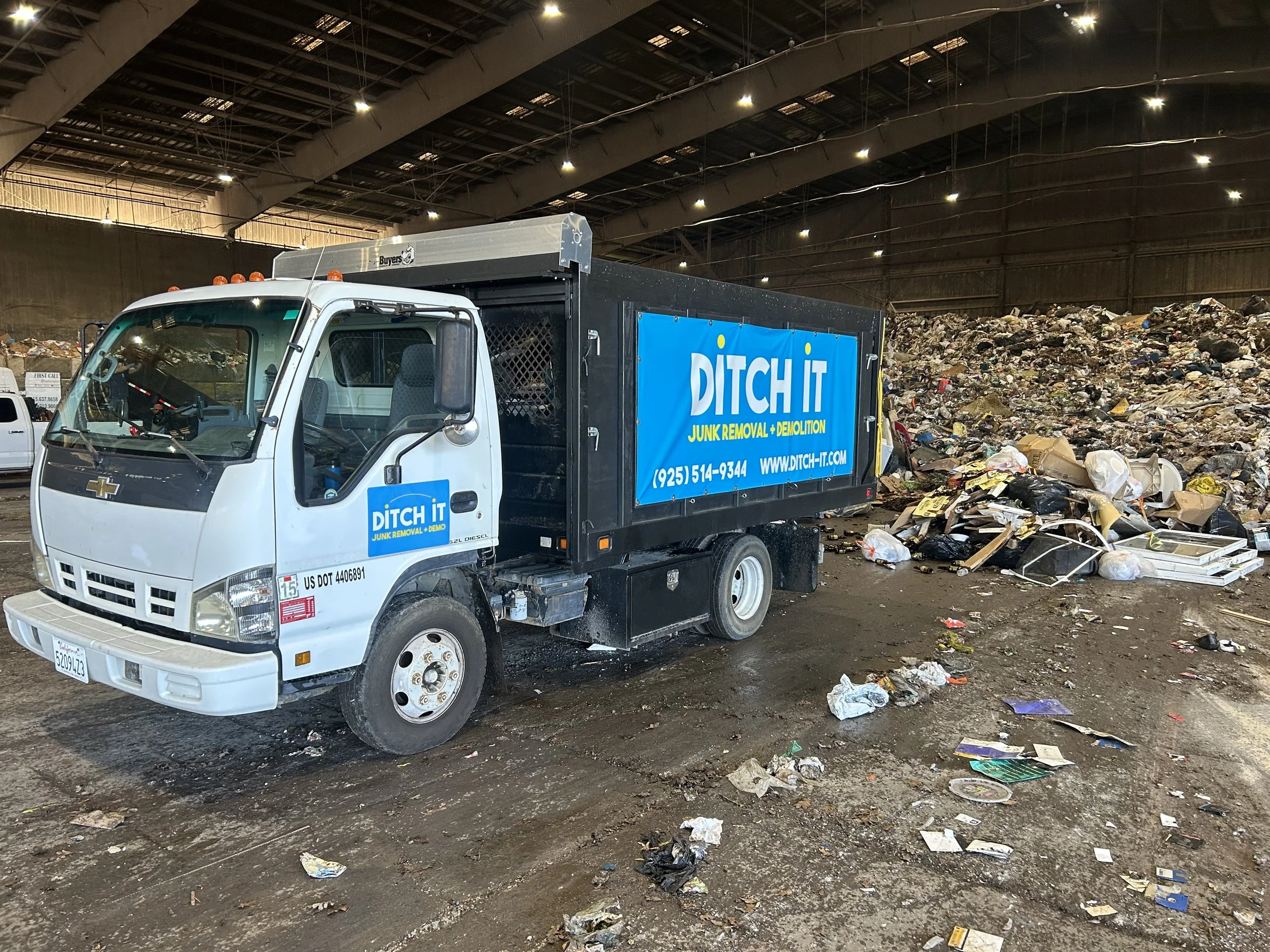 A junk removal truck labeled DITCH IT parked inside a large warehouse with a giant pile of trash and debris behind it.