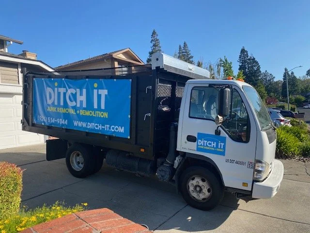 Small white truck with a blue DITCH IT banner on the side, parked in a residential driveway, with trees and houses in the background.