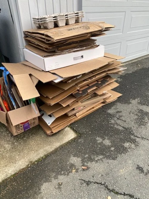 Stack of flattened cardboard boxes piled next to a trash can and a garage door, awaiting recycling collection.
