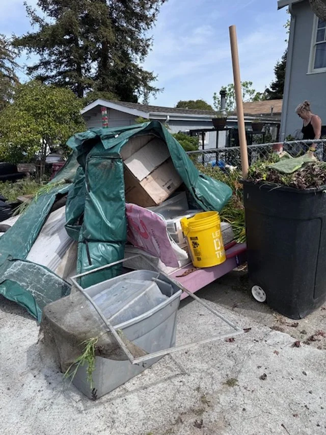 A broken green garden shed collapsed onto the ground with various gardening supplies, including a large black planter, a yellow bucket, and a gray plastic container, in a backyard with trees and a house in the background.