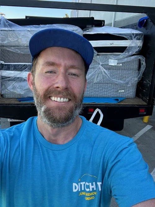 A man wearing a blue cap and blue t-shirt standing in front of a truck loaded with mattresses.