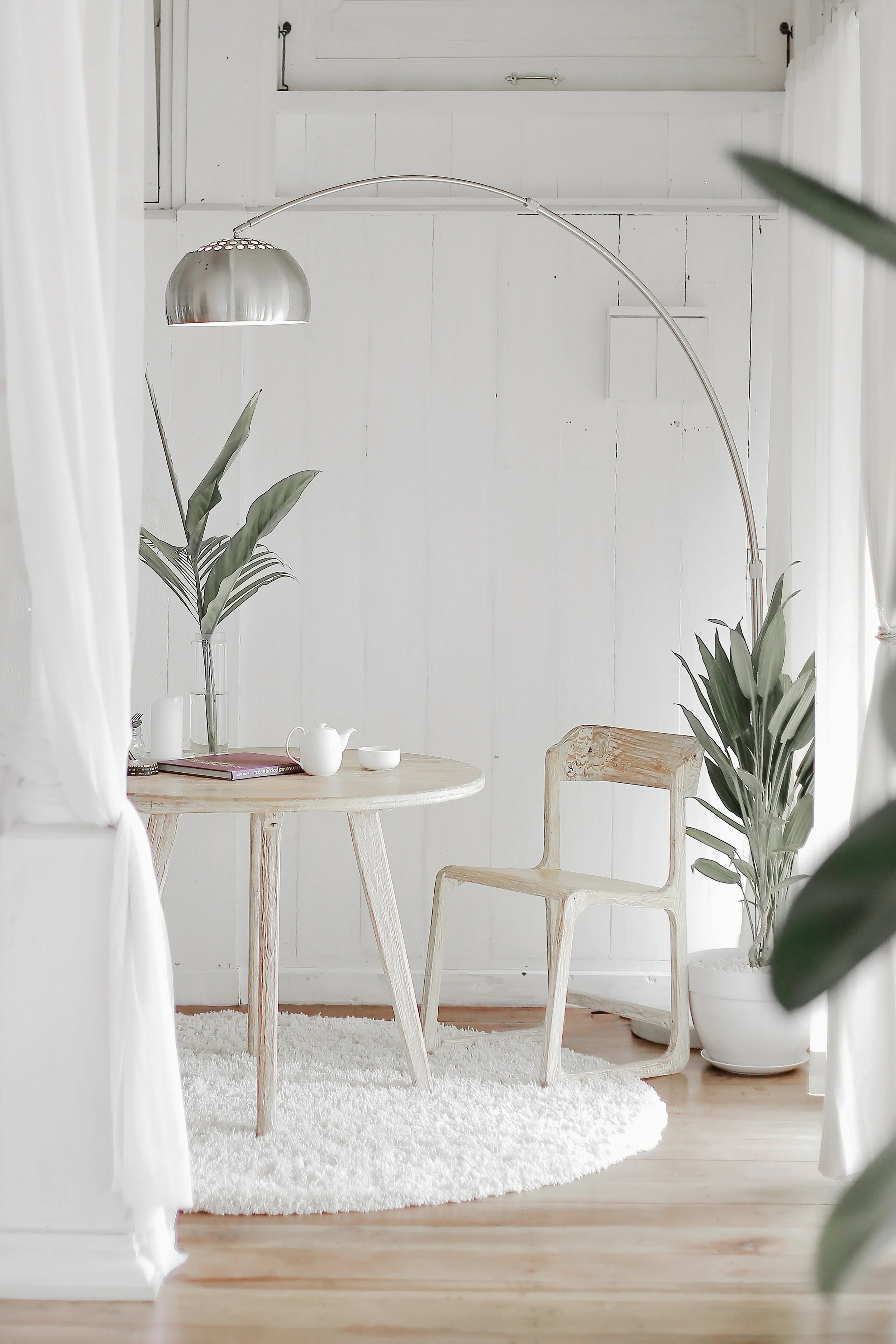 Minimalist white room with round wooden table, wooden chair, floor lamp, potted plants, and decorative items on table.