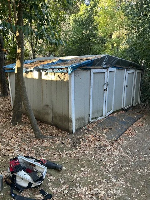 A metal storage shed with a damaged black tarp roof, located outdoors among trees, with a backpack and outdoor tools on the ground in front.