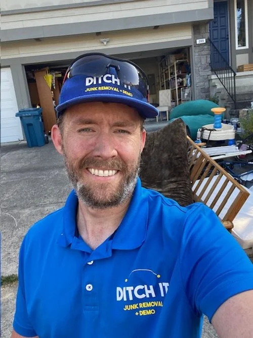 A man wearing a blue DITCH IT work shirt and a matching blue hat with the same logo, standing outside in front of a garage containing various tools and equipment, smiling at the camera.