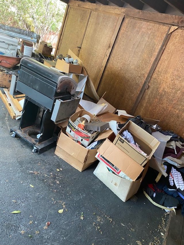 A pile of cardboard boxes, trash, and miscellaneous items in a cluttered outdoor area next to a wooden shed, with a barbecue grill and some wooden boards.
