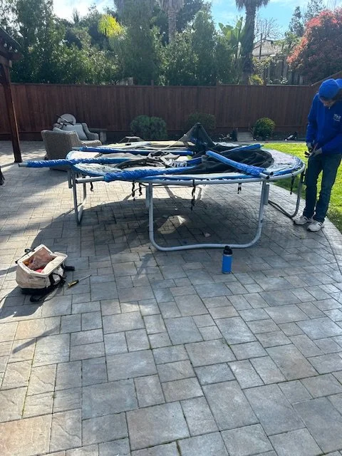 Backyard with a knocked-over trampoline, a person in a blue hoodie and cap, outdoor chairs, and gardening tools on the patio.