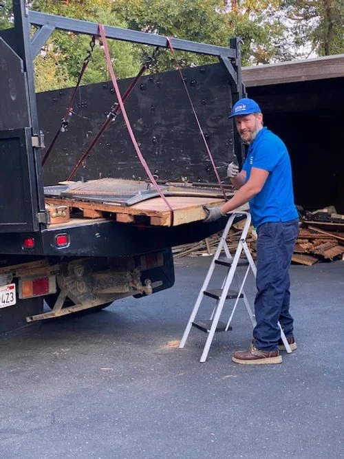 A man in a blue shirt, hard hat, and gloves standing on a step ladder, working on a pickup truck with wooden planks and red straps in the truck bed, in an outdoor area with trees in the background.