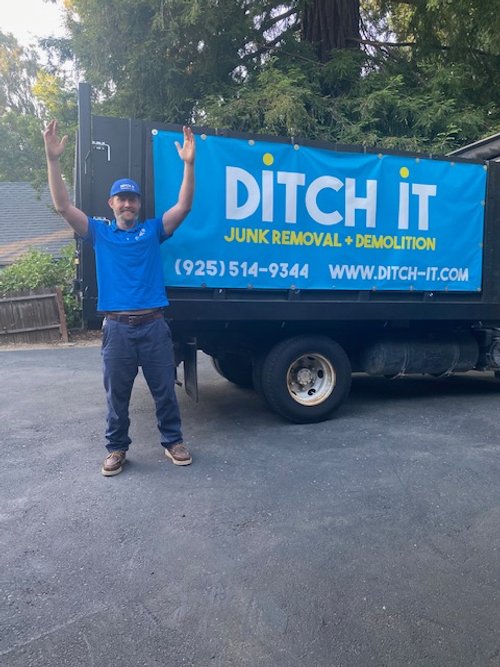 A man in a blue uniform and cap standing next to a black truck with a large blue and yellow banner that reads "DITCH IT Junk Removal + Demolition" with a phone number and website.