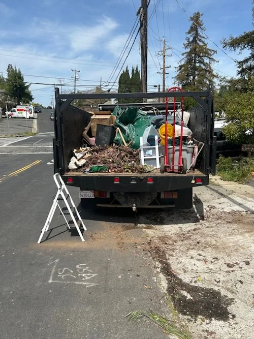 A flatbed truck filled with debris and household items, parked on a residential street. A white step ladder is set up next to the truck, and a small spot of dirt or asphalt is on the ground nearby.
