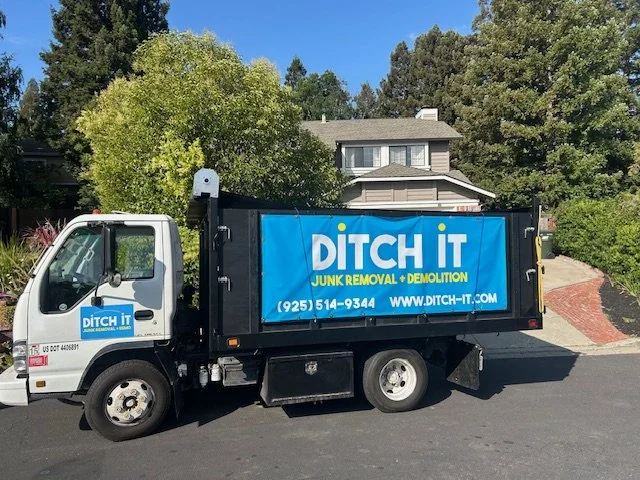 A black and white junk removal truck with the company name 'DITCH IT' on its side, parked on a paved driveway in front of a house with trees and shrubs in the background.
