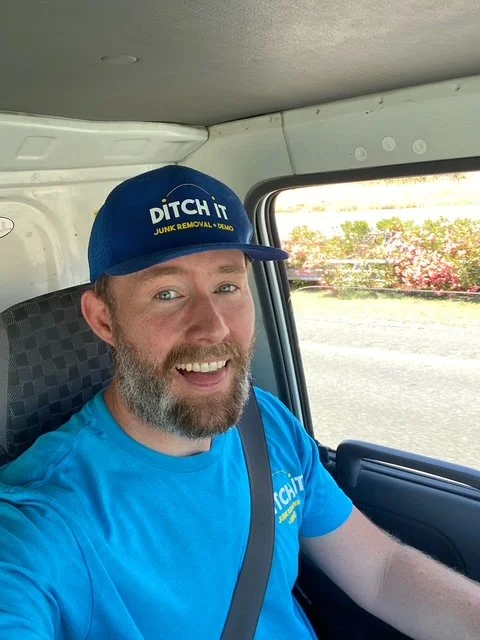 A smiling man with a beard and mustache taking a selfie inside a vehicle. He is wearing a blue cap that says "DITCH IT JUNK REMOVAL" and a blue T-shirt. The background shows a window with trees and sky outside.