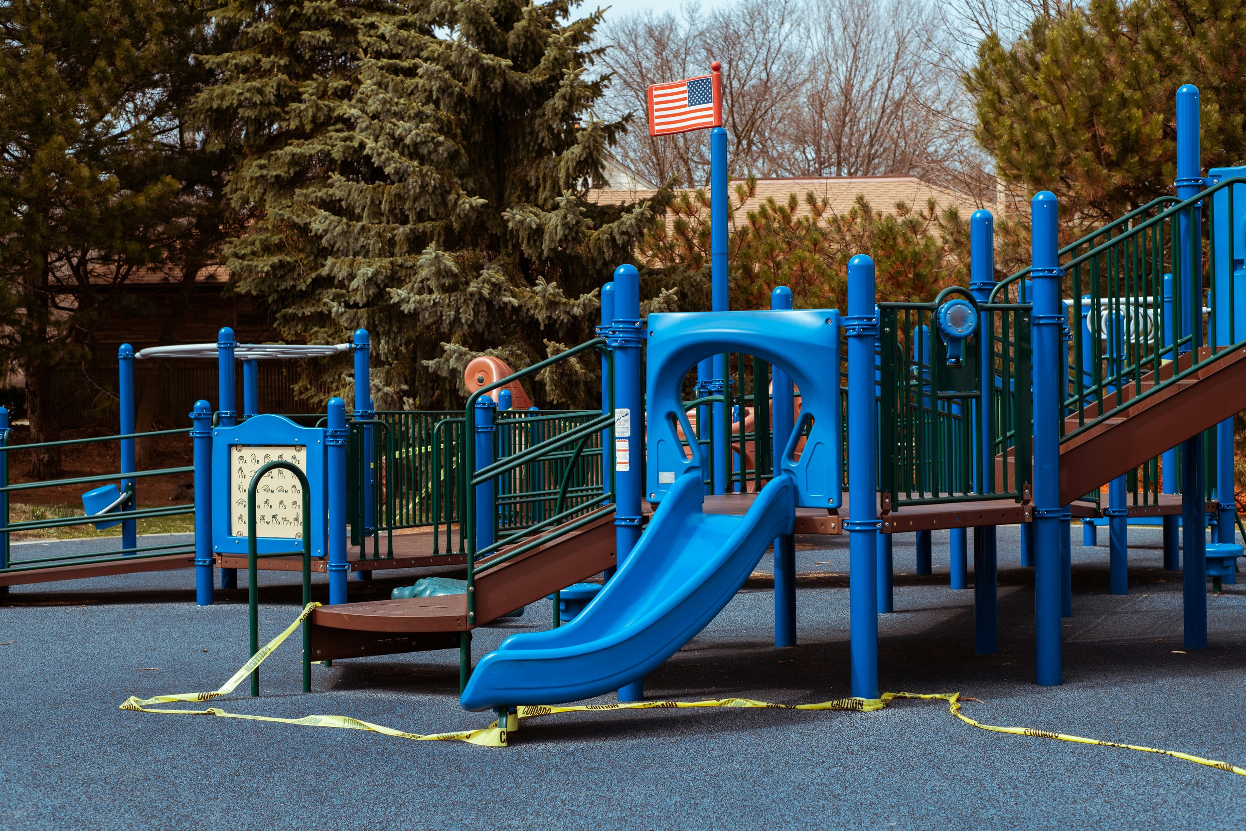 Empty playground with blue slides, climbing structures, and a caution tape around the area. American flag on a pole, large trees in the background.