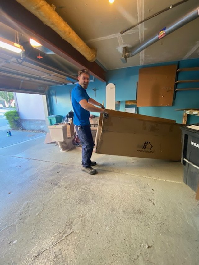 A man in a blue shirt and jeans is holding a large cardboard box inside a garage or workshop.