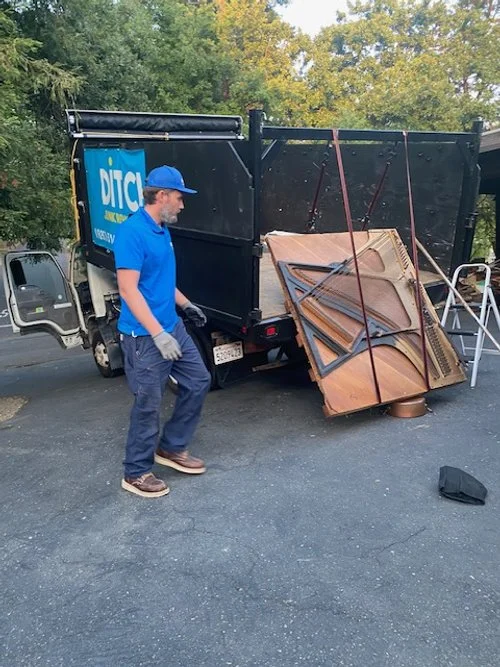 A man in a blue shirt, jeans, and a blue cap standing next to a small truck with a partially open back. There are several tables stacked and strapped to the side of the truck, and some office chairs are nearby. The truck is parked on an asphalt surface with trees in the background.
