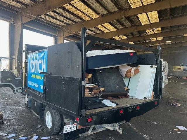 A black flatbed truck with a sign on the side that says "Junk Removal" is parked inside a large, industrial warehouse. The truck's bed is loaded with discarded furniture, boxes, and debris.