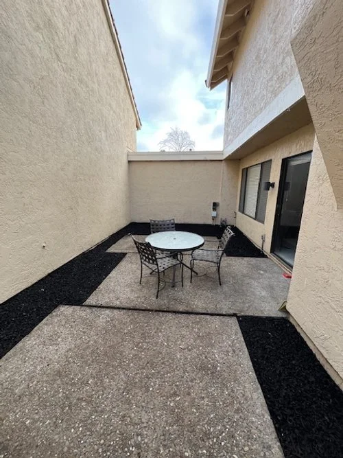 Small outdoor patio with a round glass table and three metal chairs, surrounded by beige stucco walls and a pebbled concrete floor, with patches of black mulch at the edges.